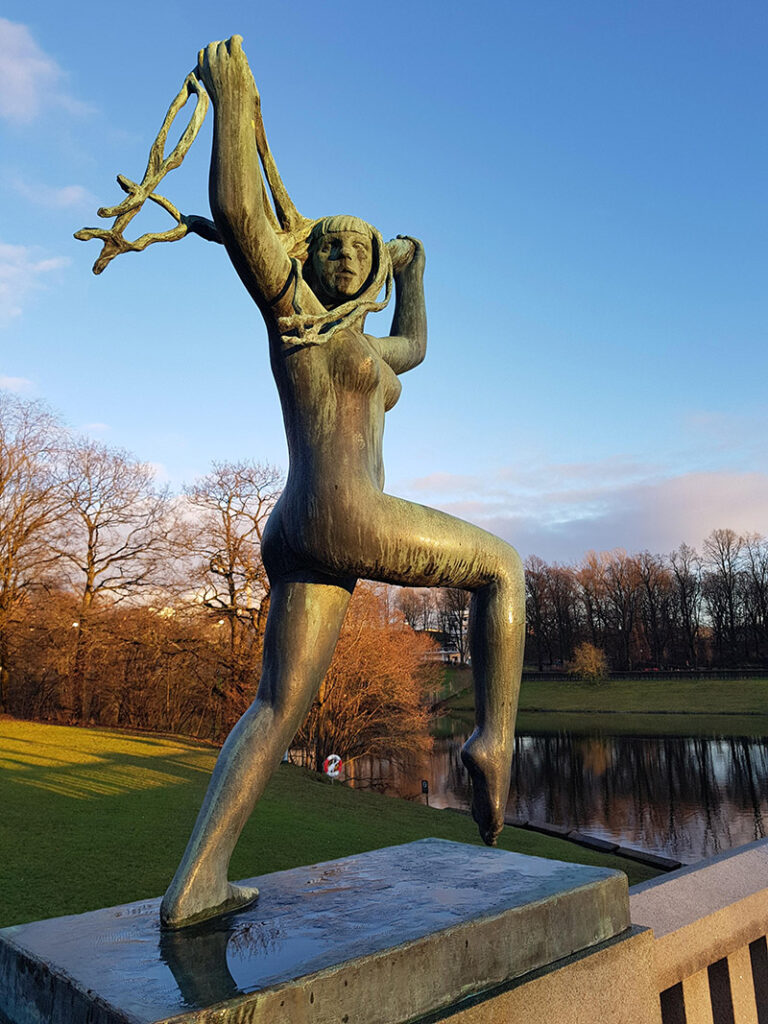 Vigeland statue av jente som springer med flagrende hår. Vigelandsparken i høstfarger i bakgrunnen.