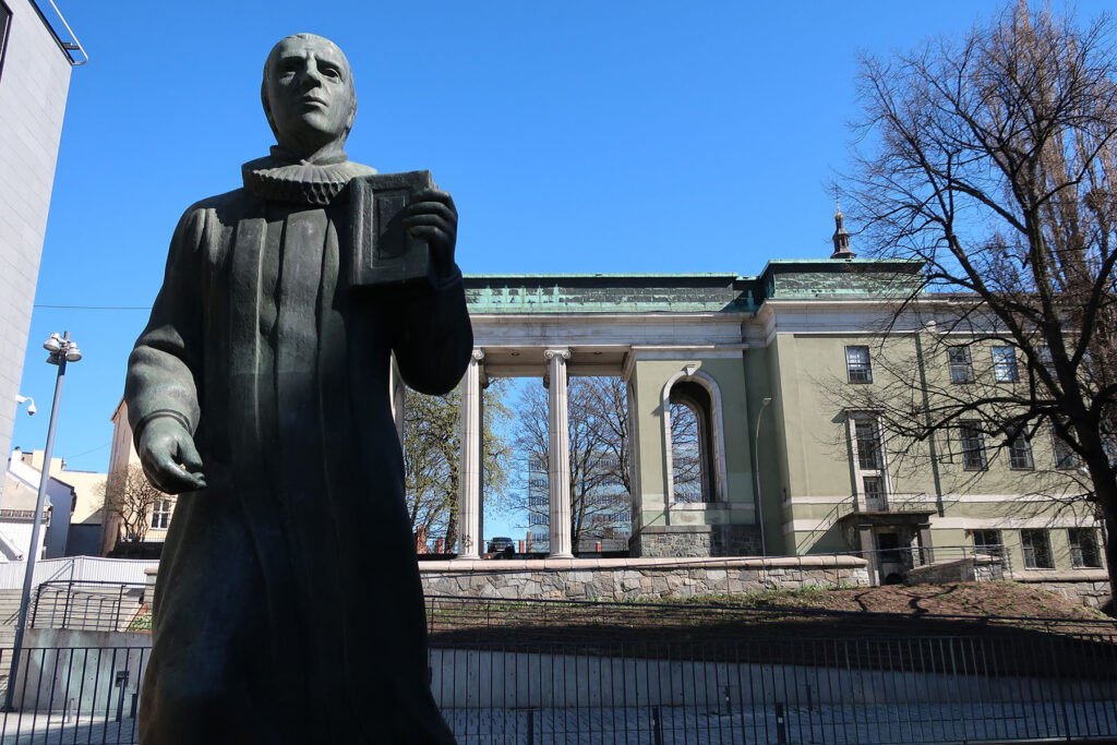 Helt i framgrunnen står en statue med prestekrage og bibel. I bakgrunnen en lys grønn bygning med høye søyler. 